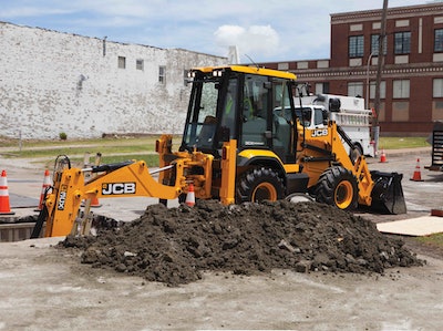 JCB 3CX Compact backhoe digging in dirt trench beneath concrete in urban area