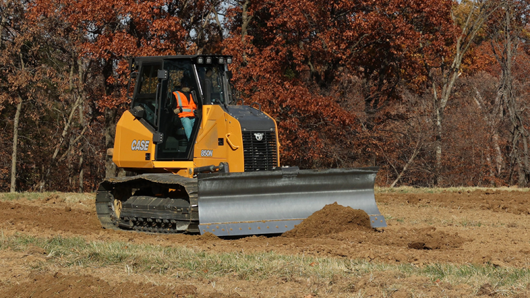 Yellow CASE bulldozer with blade on a dirt field, autumn trees in the background; a worker in the cab wears an orange safety vest (construction site)