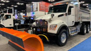 Dump truck with an oversized orange snow plow on display at a trade show, white cab and chrome accents visible indoors.