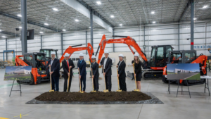 Group of professionals in hard hats shovel dirt at a groundbreaking ceremony inside a warehouse, with orange excavators in the background.