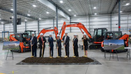 Group of professionals in hard hats shovel dirt at a groundbreaking ceremony inside a warehouse, with orange excavators in the background.