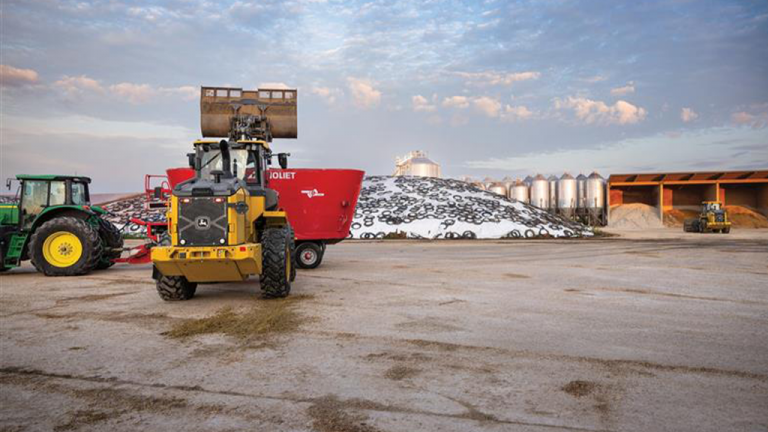 Industrial farm yard with a yellow front-end loader and a green tractor beside a red feed wagon, near a large pile of material and silos in the background.