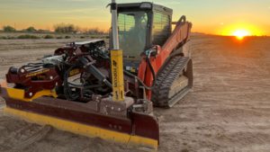 Kubota compact track loader with a wide grading blade attached, working on a dusty construction site at sunset.