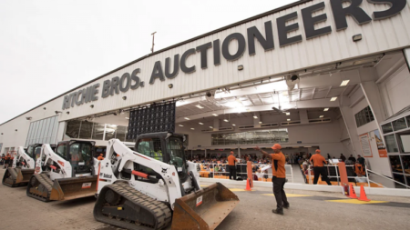 Bobcat skid-steer loaders parked outside a large Ritchie Bros. Auctioneers warehouse as staff in orange shirts direct attendees.