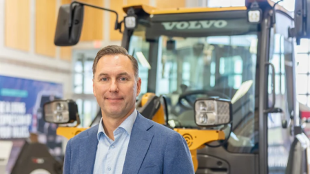 Man in a blue blazer posing in a Volvo showroom with heavy equipment in the background.