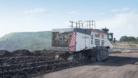 Large white-and-gray tracked mining machine on a dirt road at a quarry, with rock piles and distant hills in the background.