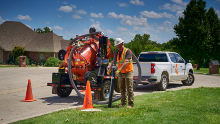 Worker in a high-visibility vest and hard hat handles a vacuum hose beside an orange septic/jetting trailer on a suburban street, cones set up nearby.