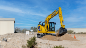 Yellow Komatsu excavator scooping dirt at an outdoor construction site under a clear blue sky, with a dirt mound and orange safety cones nearby.