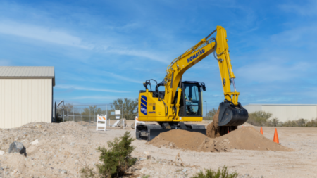 Yellow Komatsu excavator scooping dirt at an outdoor construction site under a clear blue sky, with a dirt mound and orange safety cones nearby.