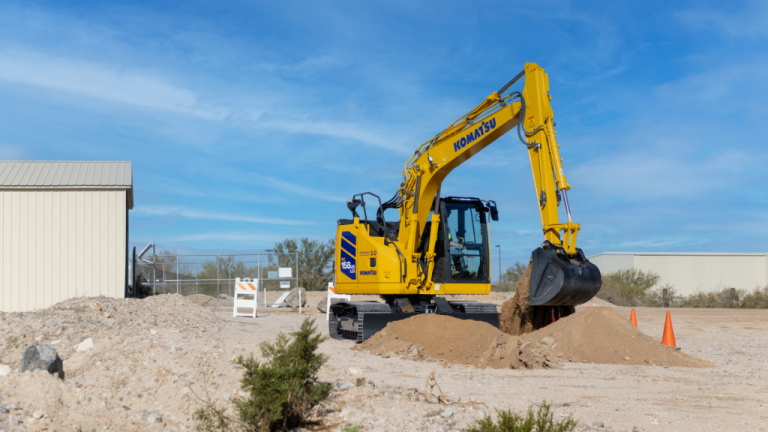 Yellow Komatsu excavator scooping dirt at an outdoor construction site under a clear blue sky, with a dirt mound and orange safety cones nearby.