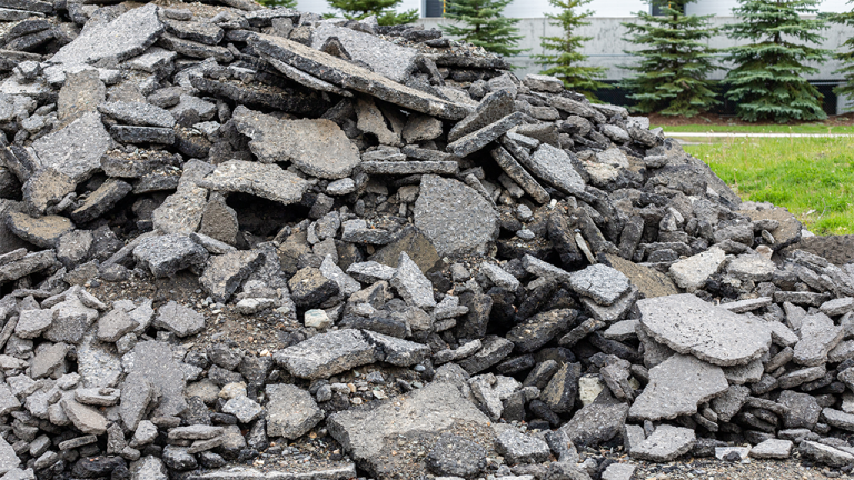 Large pile of gray crushed concrete and asphalt rubble outdoors, with grass and trees in the background.