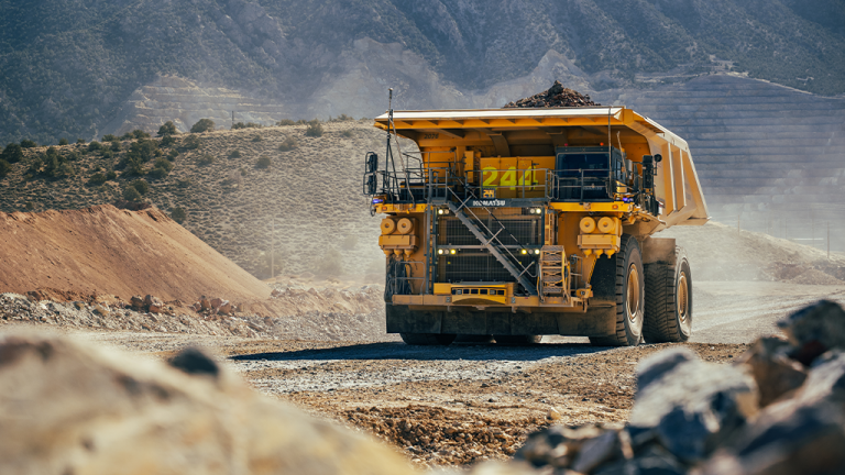 Yellow mining dump truck drives along a dusty gravel road at a quarry, mountains in the background