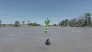 Overhead wire across a wide river with a bright green handheld device clamped to the line and a black weighted pouch hanging below.