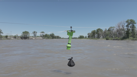 Overhead wire across a wide river with a bright green handheld device clamped to the line and a black weighted pouch hanging below.