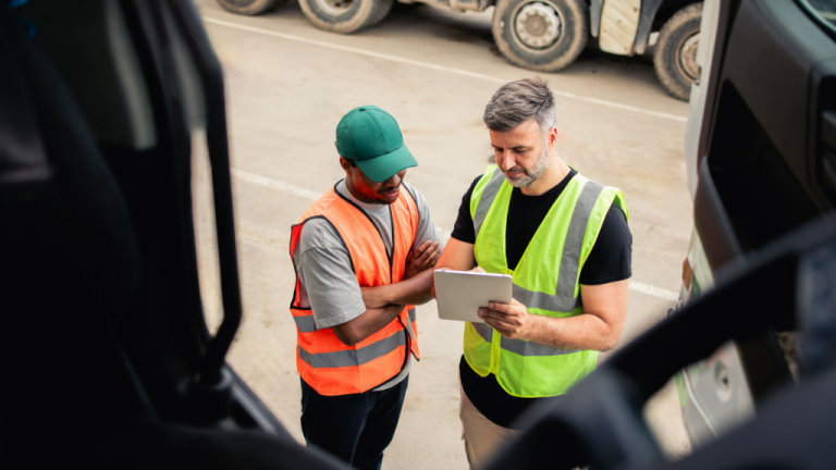 Two workers in reflective vests stand beside a truck, reviewing information on a tablet together.