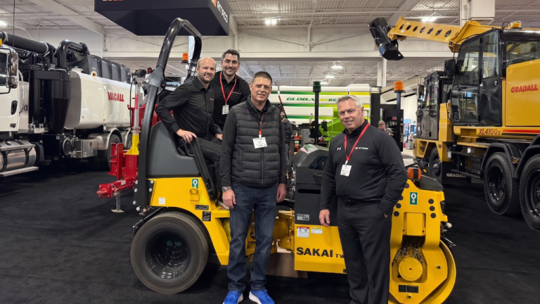 Four men wearing name badges pose beside a yellow Sakai compact roller at a construction equipment trade show, with large machinery on display in the background.