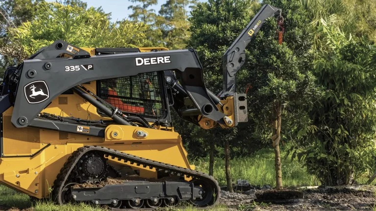 John Deere 335P tracked skid steer with a hydraulic grapple arm clearing brush near trees in a grassy area