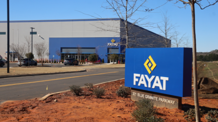 Entrance to a Fayat facility on a sunny day, with a large blue sign and yellow logo in the foreground and a blue-accented building in the background, with cars in the parking lot nearby.