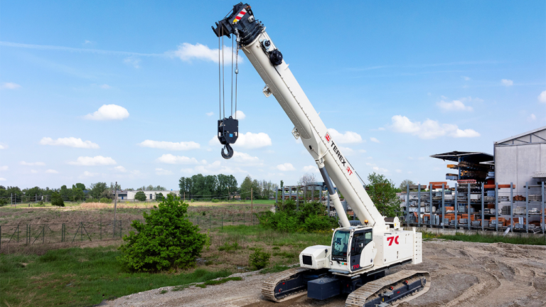 White crawler crane with an extended boom and hanging hook at a construction yard under a bright blue sky.