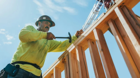 Construction worker in a hard hat and neon shirt hammering wooden framing at a building site under a blue sky