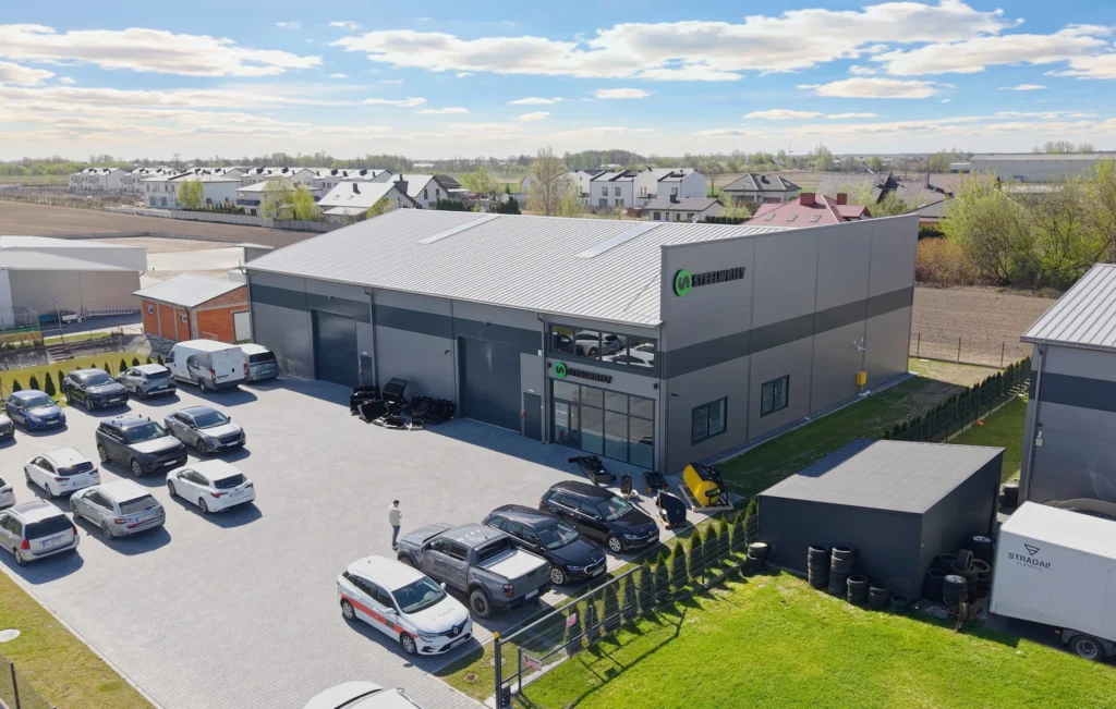 Aerial view of a gray industrial building with a metal roof and a green-and-black company logo on the corner, surrounded by a large parking lot filled with cars and a few delivery vans/ trucks.