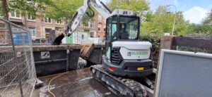 Compact excavator loading dirt into a large skip at an urban construction site, with fencing and buildings in the background.