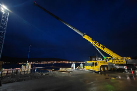 Nighttime construction site by a harbor: a large yellow mobile crane extends its boom over the area, city lights across the water in the distance.