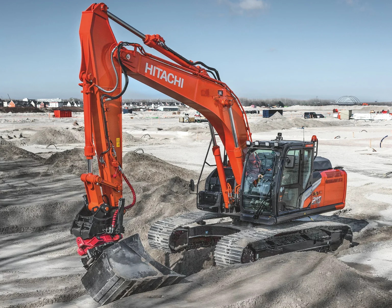 Orange Hitachi excavator digging into a sandy construction site with a long arm and bucket in the foreground