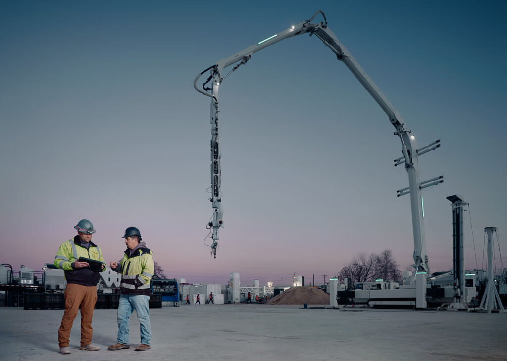 Two construction workers in hard hats and high‑visibility jackets stand on a dusty site at dusk, consulting a tablet while a giant concrete pump arm looms above them.