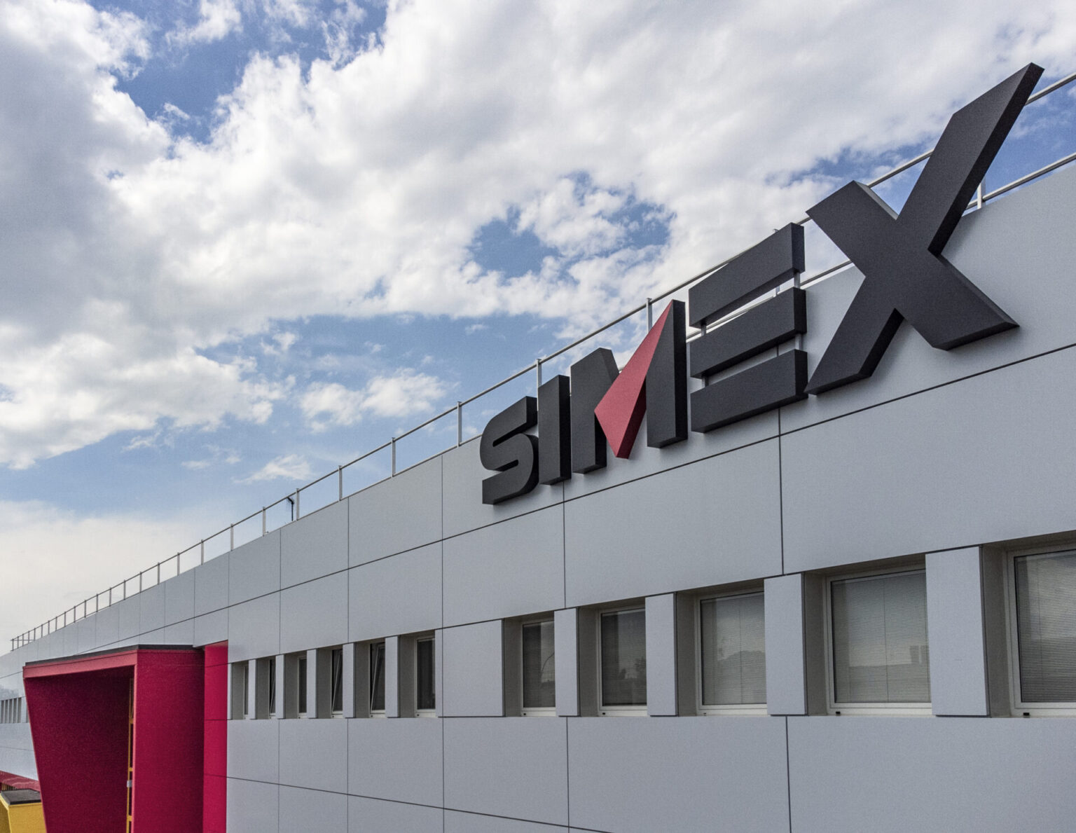 Modern office building facade with large gray logo letters and a pink vertical accent near the corner, under a partly cloudy blue sky.