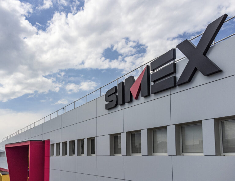 Modern office building facade with large gray logo letters and a pink vertical accent near the corner, under a partly cloudy blue sky.