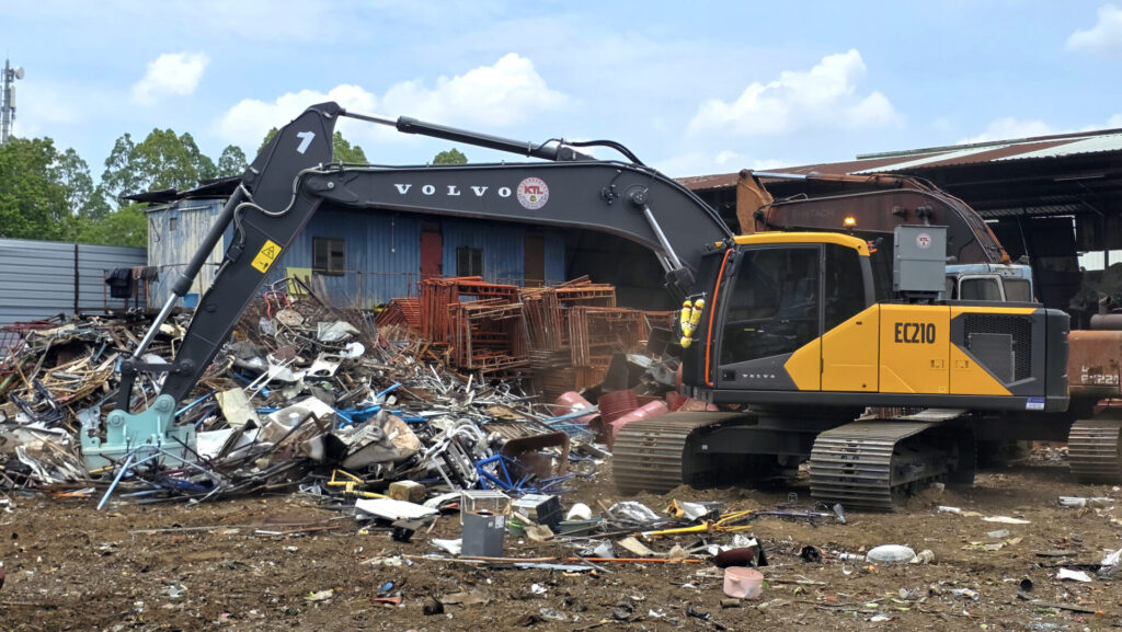 Volvo EC210 excavator at a scrap yard amid piles of metal debris and a blue building in the background.