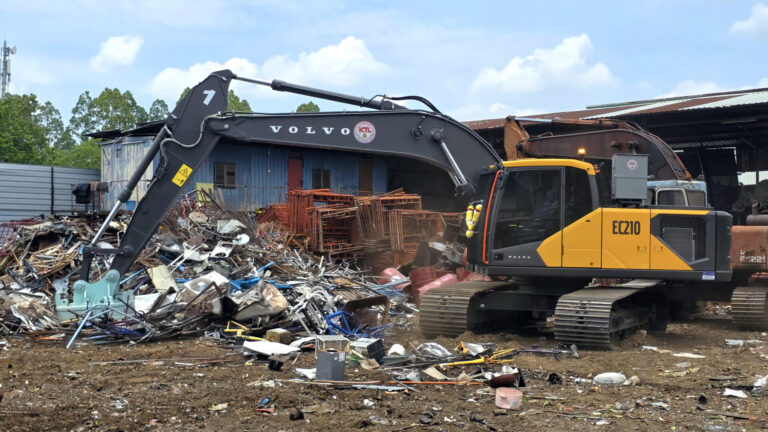 Volvo EC210 excavator at a scrap yard amid piles of metal debris and a blue building in the background.