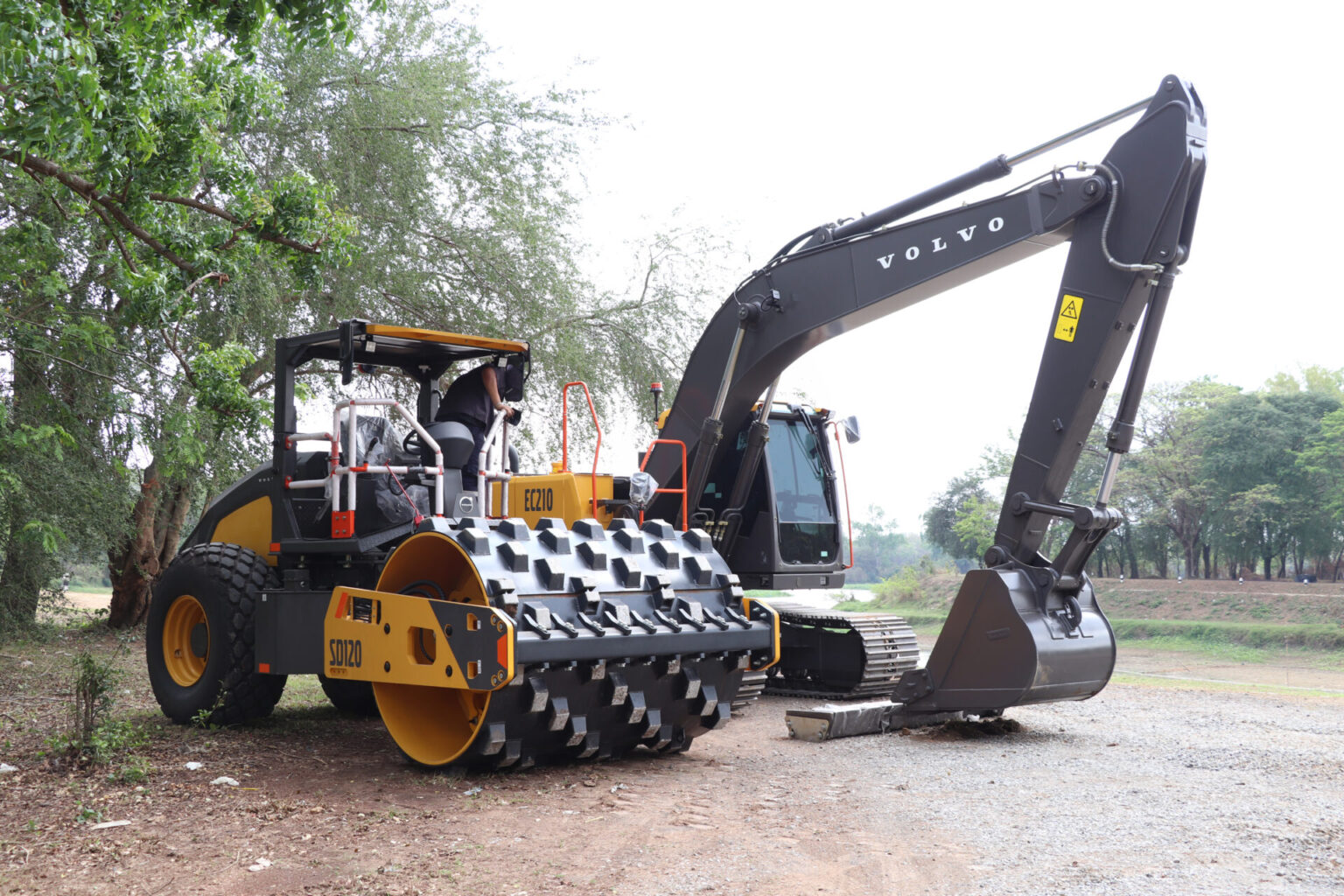 Volvo hydraulic excavator with a large front roller attachment on a dirt construction site, beside trees.