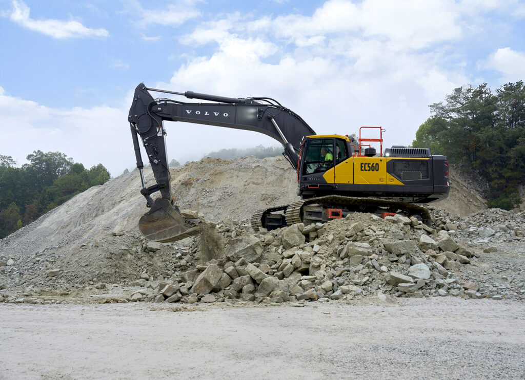 Yellow and black Volvo EC560 excavator on a rocky mound, scooping dirt with its bucket in a quarry site.