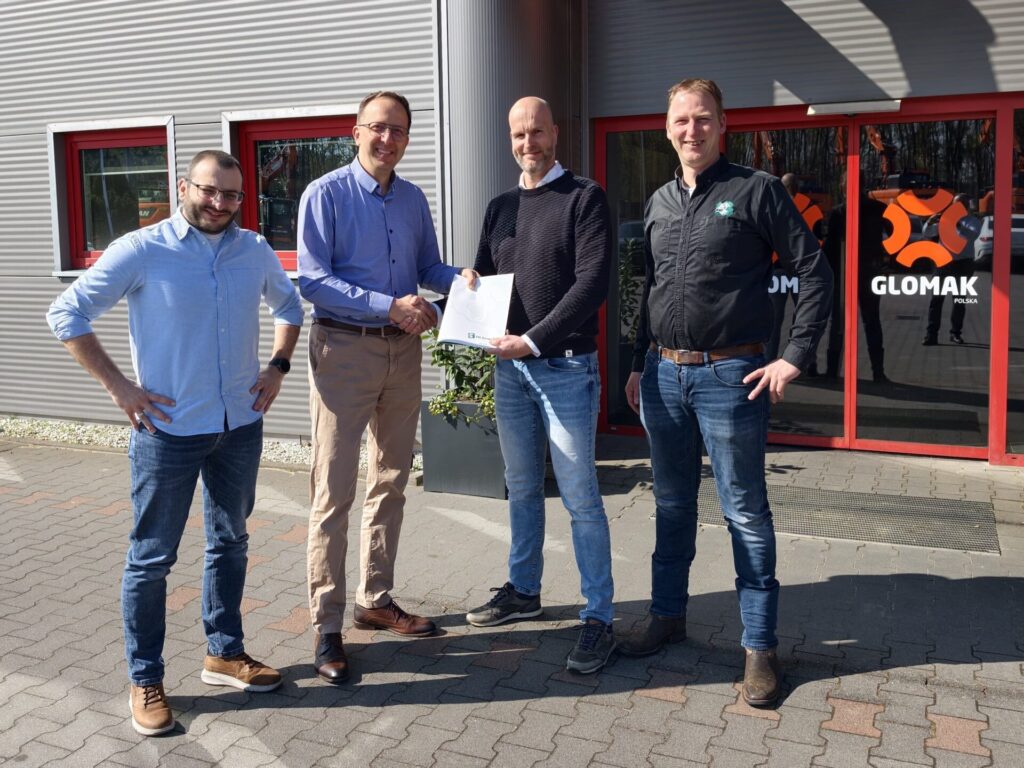 Four men stand outside a gray metal building with red-framed windows; two in the middle shake hands as one presents a certificate.
