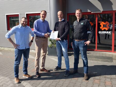 Four men stand outside a gray metal building with red-framed windows; two in the middle shake hands as one presents a certificate.