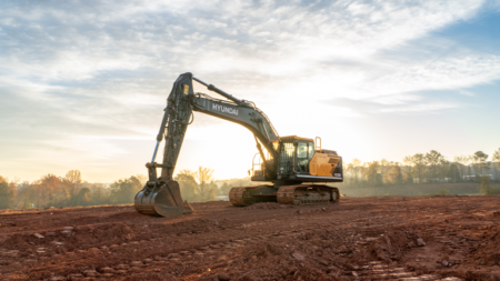 Hyundai excavator on a dirt construction site at sunset, digging into reddish soil with a tree line in the distance.