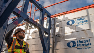 Construction worker in safety vest and hard hat stands beside a metal trench shield under blue sky, brand logos visible: United Rentals Trench Safety.