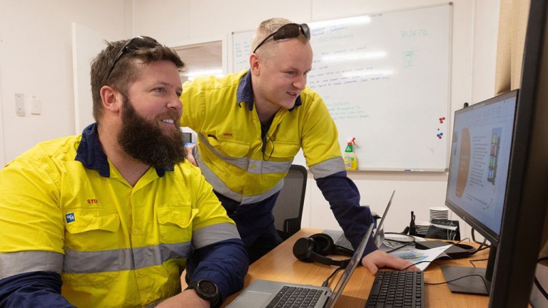 Two male workers in high-visibility yellow shirts study a computer monitor, one seated and the other standing beside him in an office.CTA