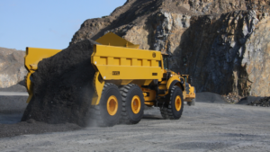 Large yellow mining dump truck unloading dark gravel at a quarry site with rocky walls in the background, wheels kicking up dust.