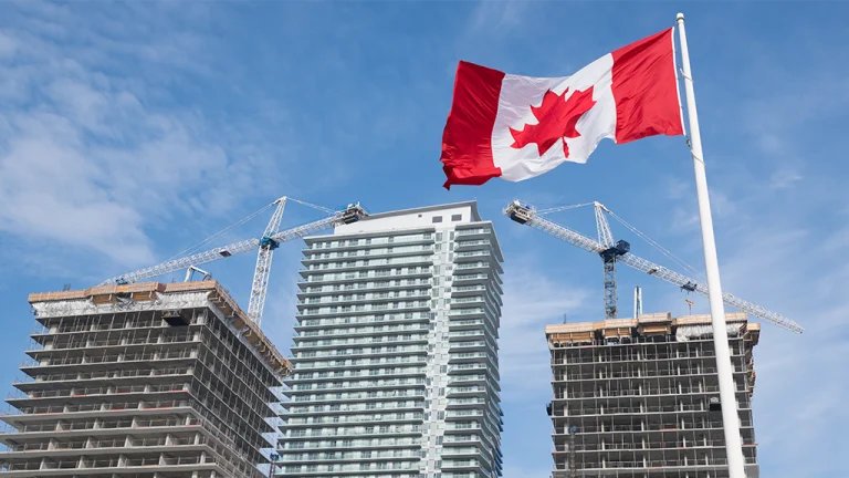 Canadian flag waves above a construction site with tall buildings and cranes against a blue sky