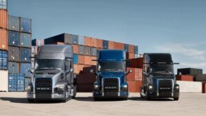 Three Mack semi-trucks parked side by side in a shipping yard, with tall stacked cargo containers in the background.
