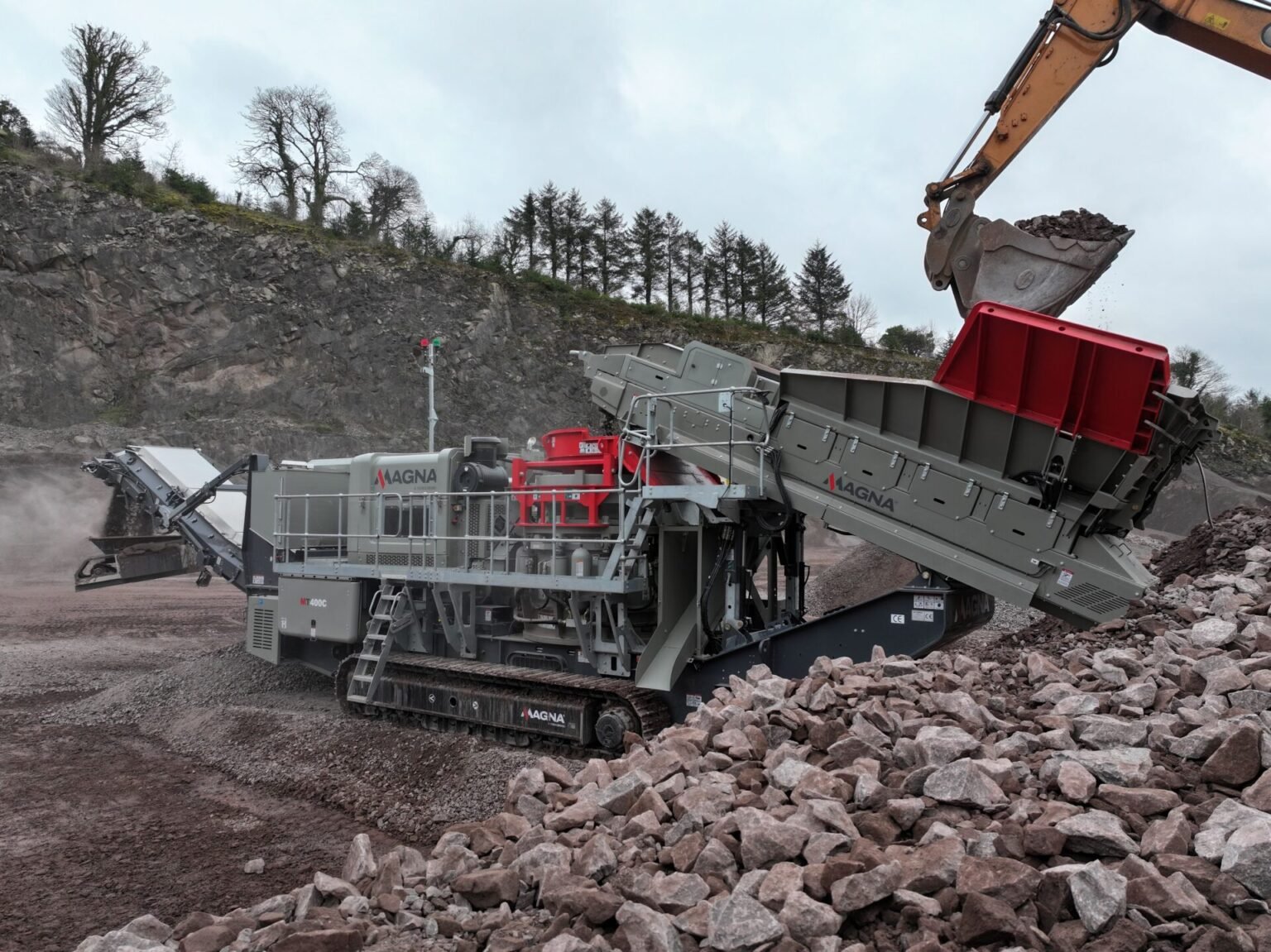 Large mobile rock crusher in a quarry receiving a load of rocks from an excavator bucket into a hopper/crusher.