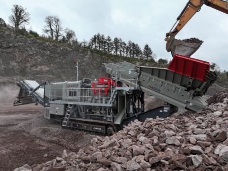 Large mobile rock crusher in a quarry receiving a load of rocks from an excavator bucket into a hopper/crusher.
