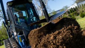1778712460_63580_en_18b44_65699_bluediamond-extreme-duty-augerpng - Machines Wiki Bulldozer digging soil, front blade raised, creating a large dirt pile in a sunny yard.