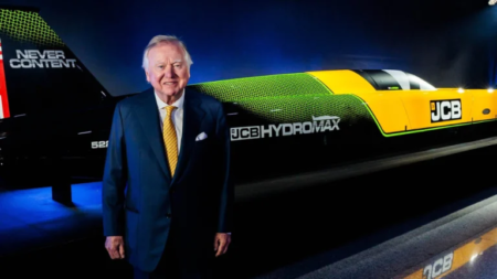 A well-dressed man in a blue suit stands beside a large yellow-and-green JCB Hydromax train showcased on a dark stage.