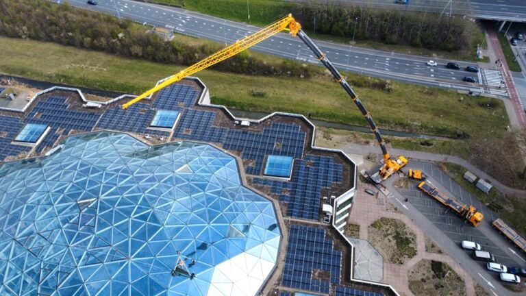 Aerial view of a glass-domed building with a large yellow crane extending over solar panels on the roof.