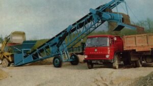 Blue inclined conveyor unloading gravel into a red dump truck at a quarry site on a dirt lot.