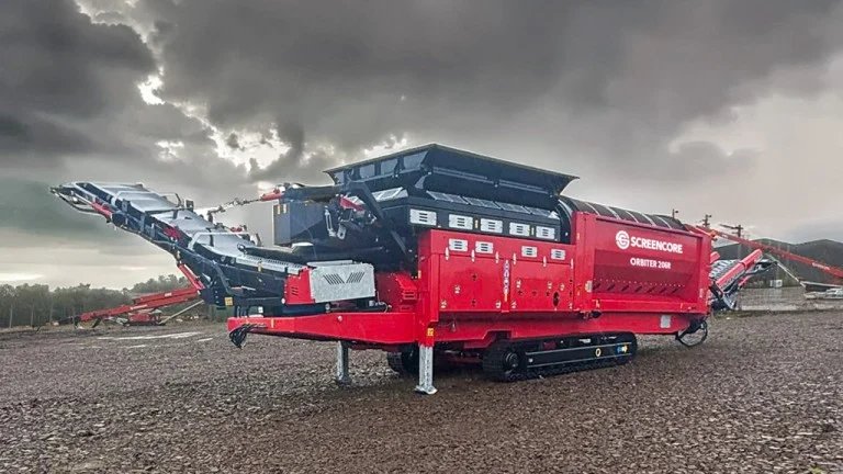 Red mobile screening plant on a gravel lot with extended conveyors under a cloudy sky.